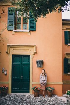 a woman sitting in front of an orange building with green shutters on the windows