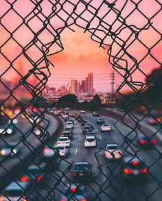 a view through a chain link fence looking at traffic on the freeway with buildings in the background