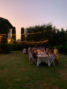 a group of people sitting around a table outside at night with lights strung across the lawn