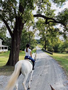 a woman riding on the back of a white horse down a dirt road next to a tree