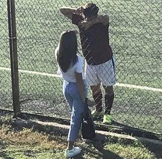 two people standing next to each other near a fence and grass area with a tennis court in the background