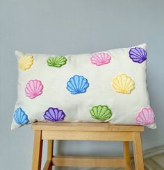 a white pillow with colorful seashells on it sitting on a wooden stool next to a gray wall