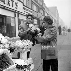 an old black and white photo of two women standing on the sidewalk with flowers in front of a flower shop