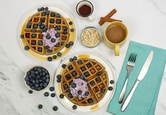 two waffles with blueberries and other toppings on plates next to silverware