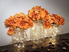 three mason jars filled with orange flowers on top of a wooden table next to string lights