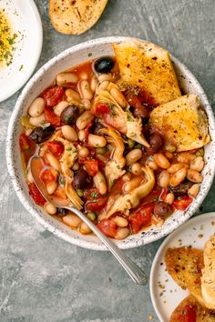 a white bowl filled with beans and vegetables next to two plates of bread on the side