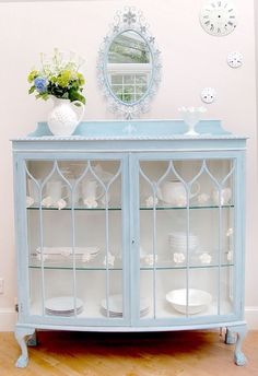 a blue china cabinet with glass doors and plates on it's sides, in front of a wall clock