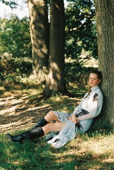 a woman sitting on the ground next to a tree wearing boots and a white dress