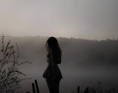 a woman standing in front of a body of water on a foggy day with her back to the camera