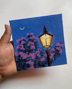 a hand holding up a small painting of a street light and flowers in the background