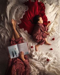 two people laying on a bed with red sheets and pillows, one reading a book