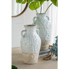 two white vases sitting on top of a wooden table next to a green plant