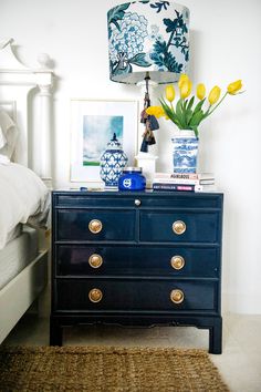 a blue dresser with yellow tulips in front of a white bed and lamp