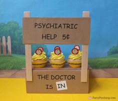 three cupcakes with yellow frosting sitting on top of a cardboard sign that says psychic help 56 the doctor is in