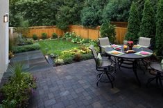 an outdoor dining area with brick pavers and wooden fenced in trees, shrubs, and flowers