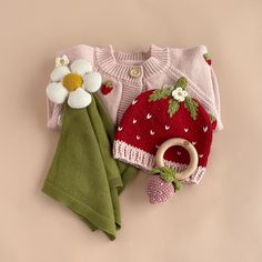 a knitted strawberry sweater and matching hat are displayed on a pink surface with a green cloth