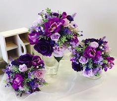 three vases filled with purple flowers sitting on top of a white cloth covered table