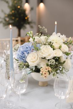 a white table with blue and white flowers in a bowl next to some wine glasses