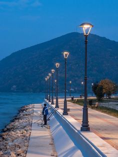 a person sitting on a bench next to the water at night with street lights and mountains in the background