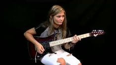 a woman is playing an electric guitar while wearing white pants and black shirt with her legs crossed