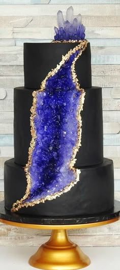 a black cake with purple and gold decorations on it's tiered layer, topped with a large blue druzby rock