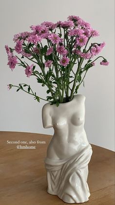 a white vase with pink flowers in it on a wooden table next to a wall