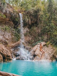 a woman hanging from a rope in front of a waterfall with blue water and trees