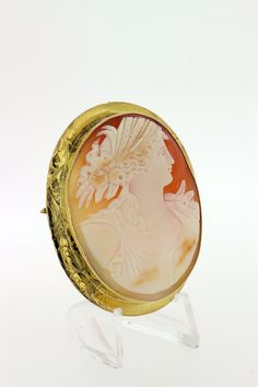 This delightful cameo brooch is made in 14K yellow gold and oval cut cut piece with a Greek motif with a female figure. She's detailed with braided hair, feathers, and roses. The brooch is accented with an engraved bezel and migraine edge. It can also be worn as a necklace too--has a toggle that can flip up or down. The entire piece measures 57mm long, 46.31mm wide, 11.33mm tall, and weighs 10.7DWT. B55KIPPN --Please reference our policy for more details before purchase-- For International order Luxury Oval Cameo Brooch, Heirloom Oval Brooches With Intaglio, Luxury Oval Intaglio Brooches, Yellow Gold Oval Cameo Brooch, Gold Oval Carved Brooches, Oval Carved Gold Brooches, Luxury Oval Engraved Brooches, Hair Feathers, Convertible Necklace