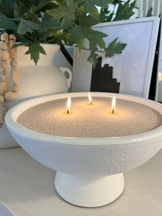 three lit candles in a white bowl on a table next to vases with green leaves