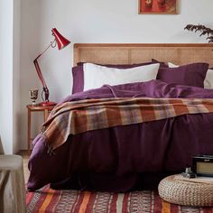 a bed with purple sheets and pillows in a bedroom next to a table with a lamp on it