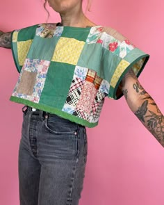 a woman with tattoos on her arm wearing a patchwork shirt and denim jeans, standing against a pink background