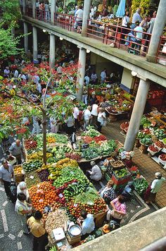 an outdoor market with lots of vegetables and people standing around it in front of the building