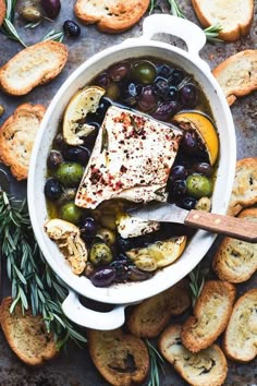 a pot filled with meat and olives next to crackers, bread and rosemary