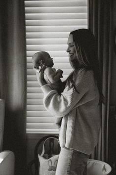 a woman holding a baby in her arms while standing next to a window with blinds