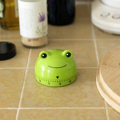 a green frog toy sitting on top of a kitchen counter next to bottles and measuring tape