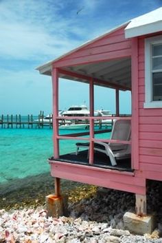 a pink house sitting on top of a rocky beach next to the ocean with boats in the water