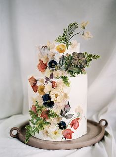 a white wedding cake with flowers and greenery on the top is sitting on a metal platter