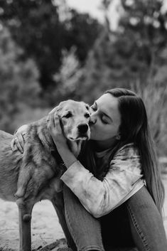 a woman is kissing her dog on the nose while sitting in front of some trees