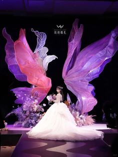 a woman in a white dress standing on a runway with purple and pink wings above her head