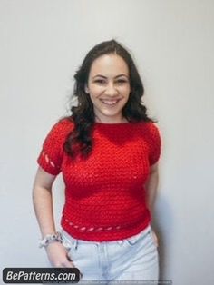 a woman standing in front of a white wall wearing a red shirt and jeans with her hands on her hips