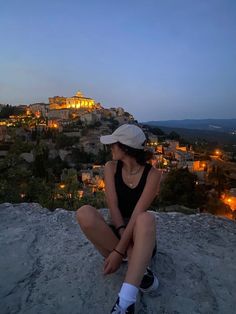 a woman sitting on top of a rock next to a town at night with lights in the background