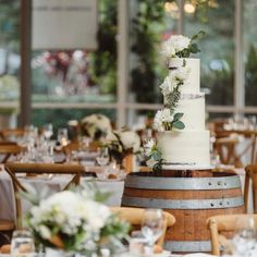 a wedding cake sitting on top of a wooden barrel in front of tables with white flowers