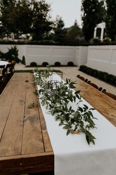 the table is set with white linens and greenery on it, along with long wooden benches