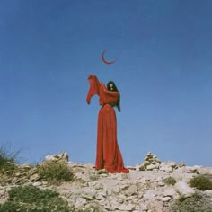 a woman in a long red dress standing on top of a rocky hill with a crescent above her head