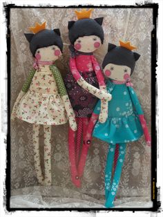 three dolls are sitting next to each other on a cloth wallpapered background, one is wearing a dress and the other has a crown