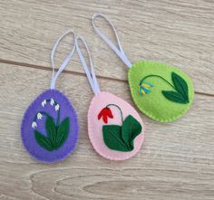 three felt ornaments with flowers on them sitting on a wooden surface, one is green and the other is pink