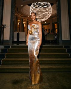 a woman in a gold and silver dress standing on some steps with a chandelier above her head