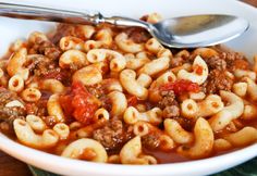 a white bowl filled with pasta and meat on top of a wooden table next to a spoon