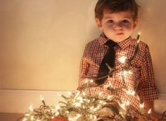 a little boy sitting on the floor next to a christmas tree with lights around him