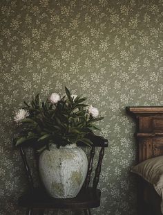 a white vase with flowers sitting on top of a wooden chair next to a bed